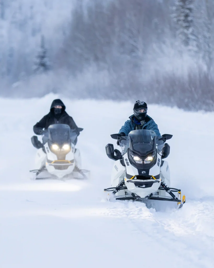 Two electric snowmobiles following a guide through a groomed winter wilderness trail.